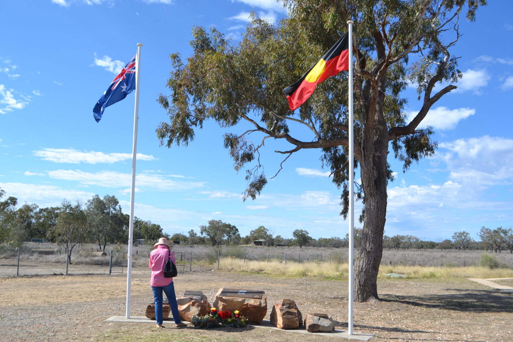 A woman in a pink shirt stands in front of a memorial between the Australian flag and the Aboriginal flag.