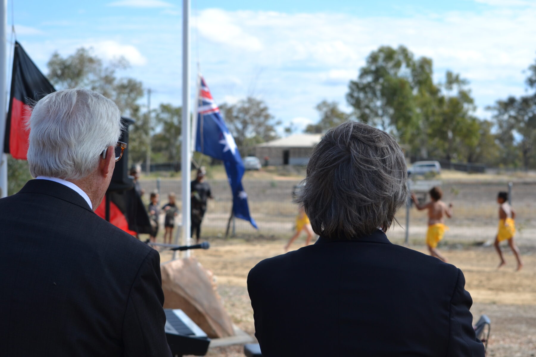 The back of two mens heads watching young children dance at a ceremony