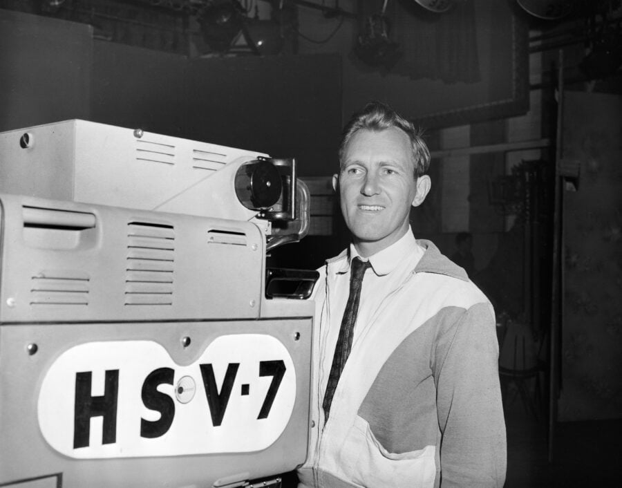 A black and white photo of a camera man standing behind a HSV 7 television camera.